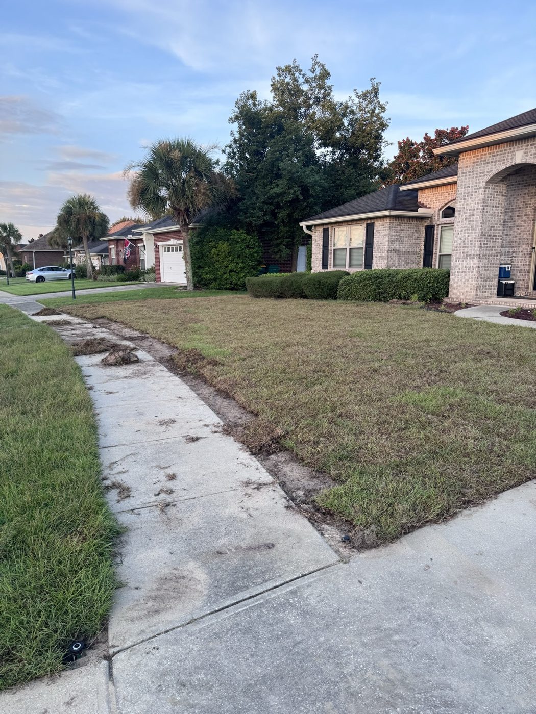 A narrow trench of bare soil dug along a concrete sidewalk, with dirt clumps on the walk and a brick house in the background.